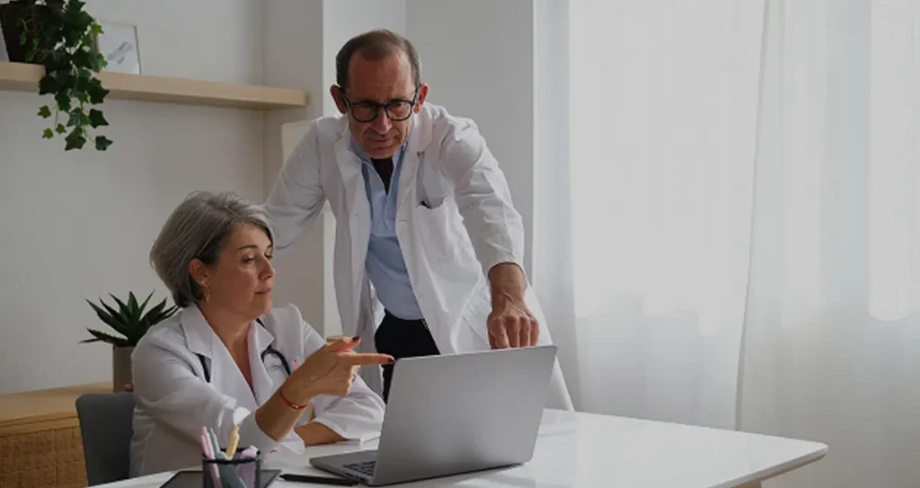 Two health care providers in white coats discuss something on a laptop in a bright office. The atmosphere is professional and collaborative.