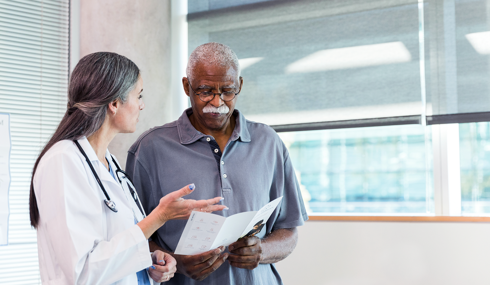 An older Black man holds medical information, which he reviews with a physician who is a woman with light skin.