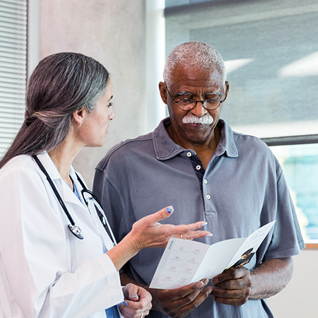 An older Black man holds medical information, which he reviews with a physician who is a woman with light skin.