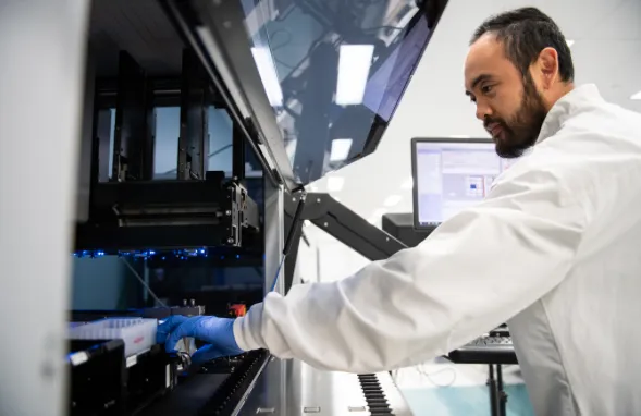 A lab worker in a lab coat and gloves operates a high-tech laboratory machine. The setting is bright and clean, emphasizing precision and focus.