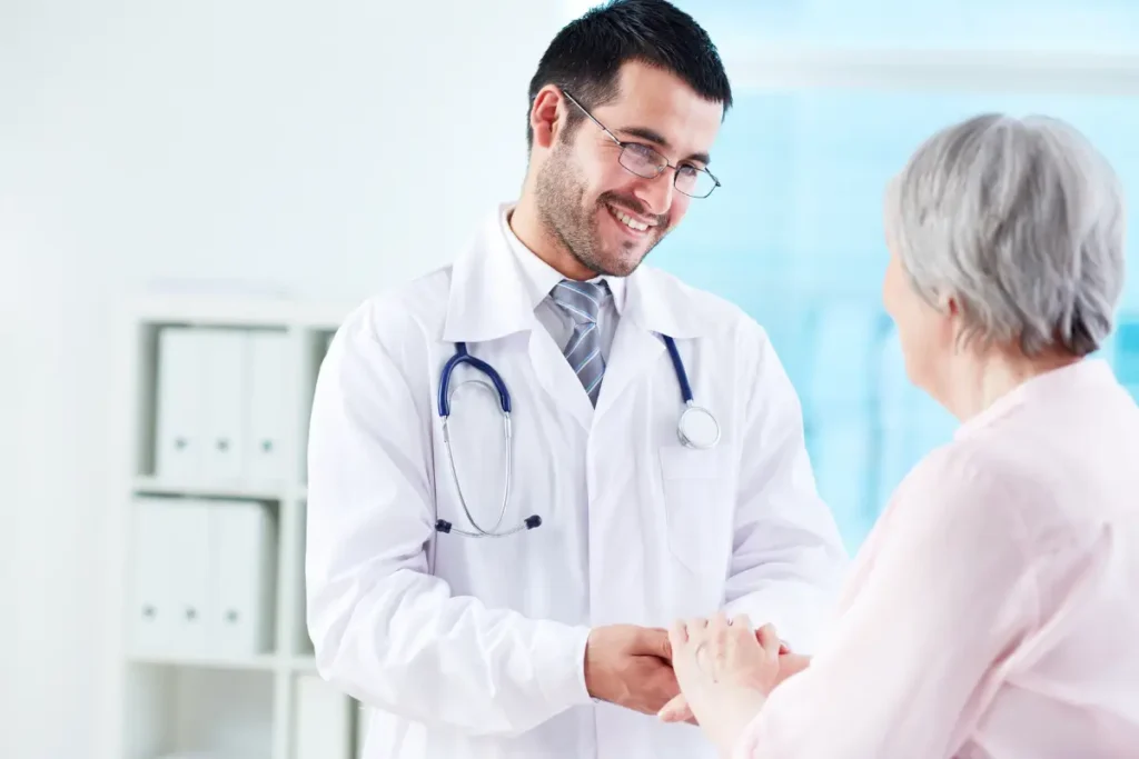 A health care provider in a white coat smiles warmly, holding an elderly woman's hands in a comforting manner. The setting is bright and clinical, conveying care.