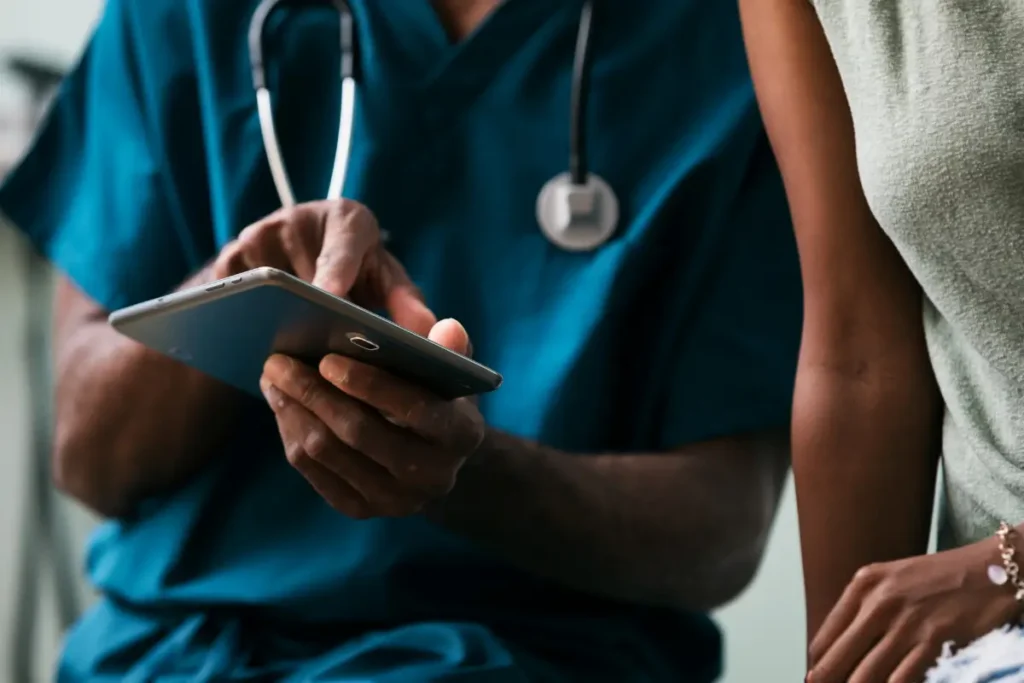 A healthcare professional in blue scrubs uses a tablet, wearing a stethoscope, while consulting with a person in casual attire. The scene is collaborative.