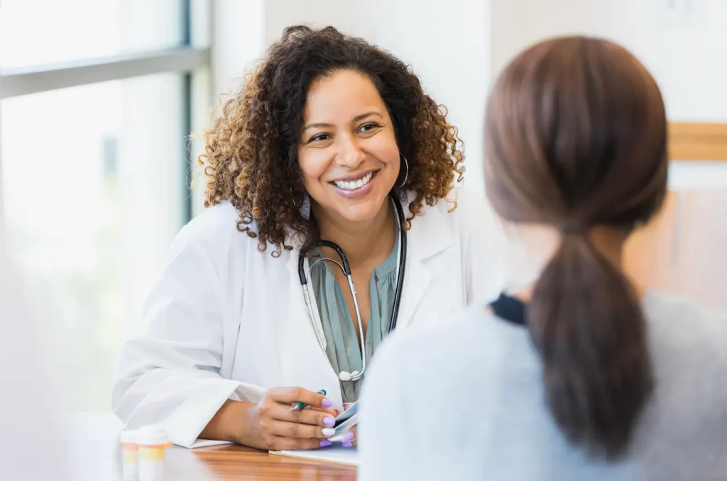 A smiling health care provider with curly hair, white coat, and a stethoscope talks to a patient. They sit across a table in a bright, welcoming room, conveying warmth and care.