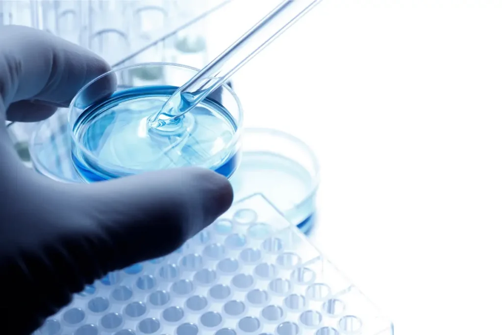 A gloved hand holds a petri dish filled with blue liquid, as a pipette dispenses fluid. Nearby are other lab dishes and a microplate, suggesting a laboratory setting.