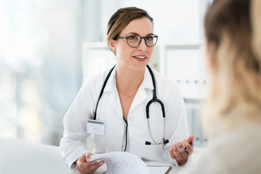 A young female health care provider with glasses and a stethoscope, smiling and holding papers, converses with a patient in a bright office. The setting feels professional and welcoming.