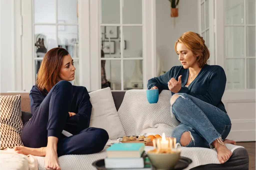 Two women sit on a cozy sofa engaged in conversation. One, holding a mug, gestures while the other listens intently. A warm, relaxed atmosphere with candles lit.