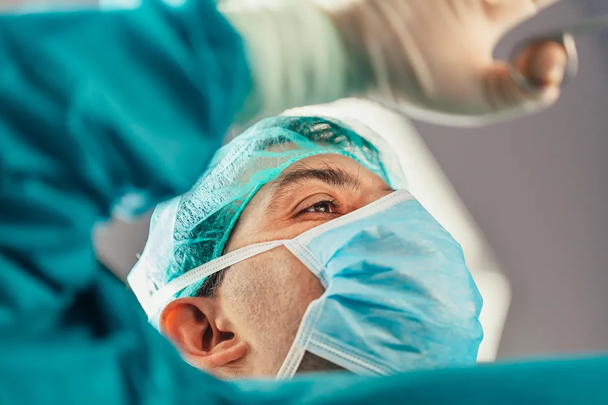 A surgeon in a blue surgical mask and cap focuses intently, with gloved hands raised. The close-up conveys concentration and precision.