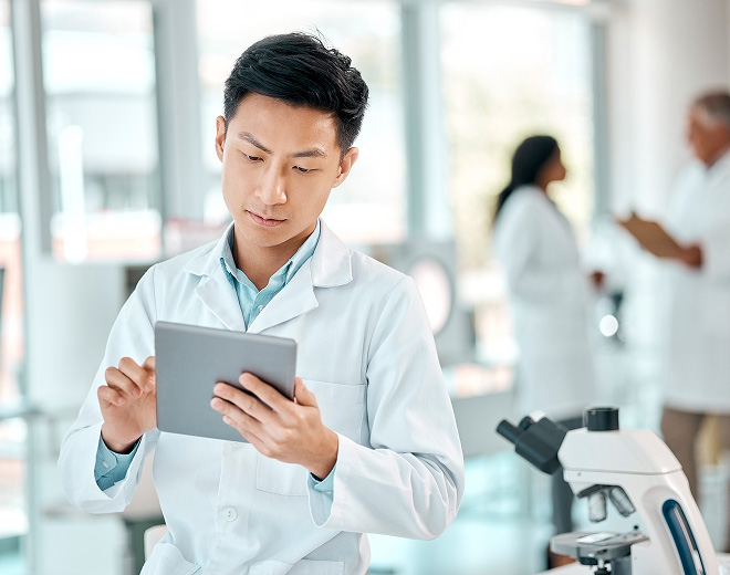 Young lab worker in a lab coat uses a tablet near a microscope. Two colleagues converse in the background. The tone is focused and professional.