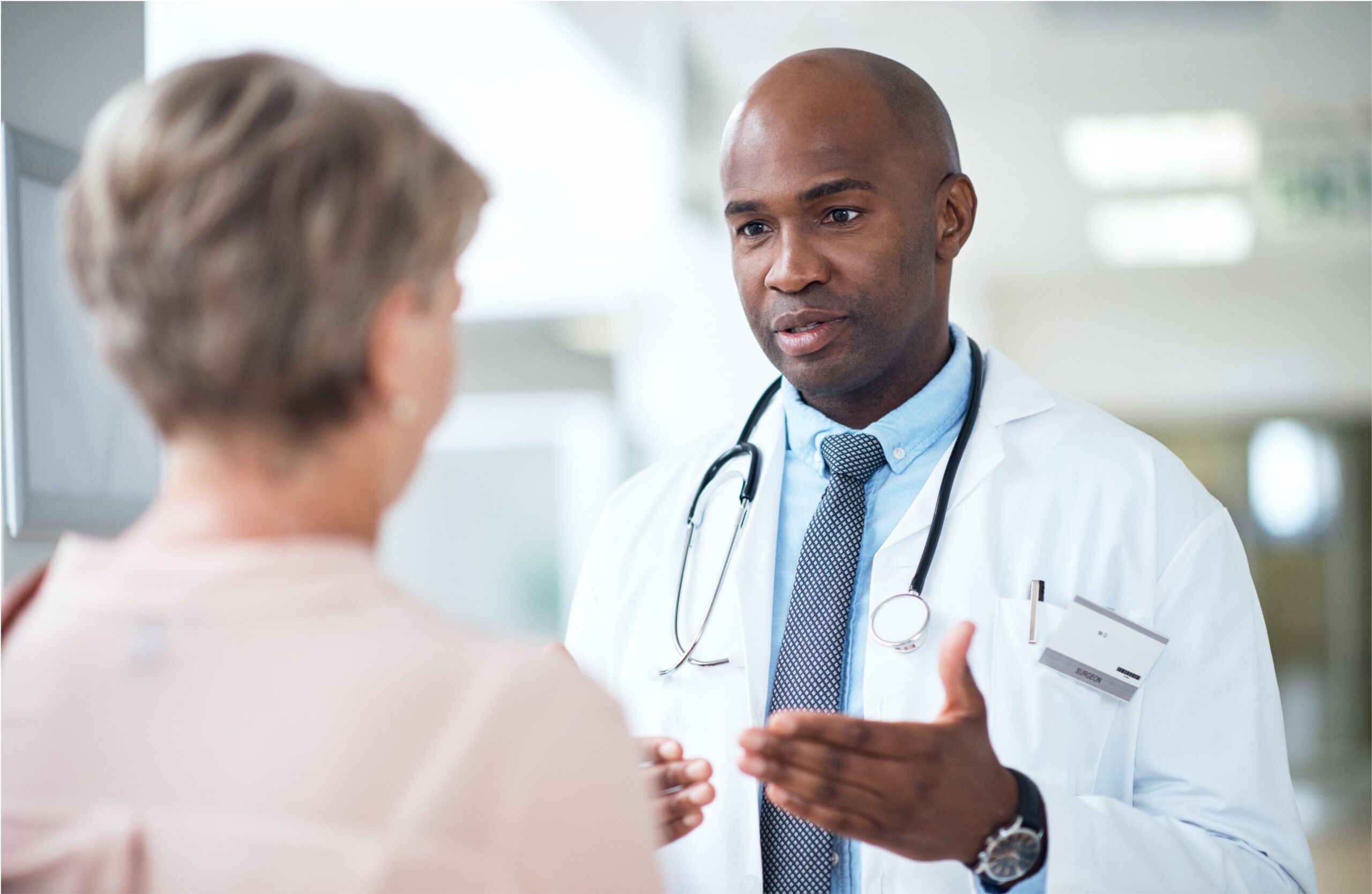 A health care provider in a white coat and stethoscope attentively talks to a patient. The setting is a bright medical office, conveying professionalism and care.