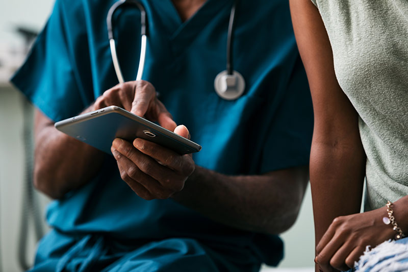 A healthcare professional in blue scrubs uses a tablet, wearing a stethoscope, while consulting with a person in casual attire. The scene is collaborative.