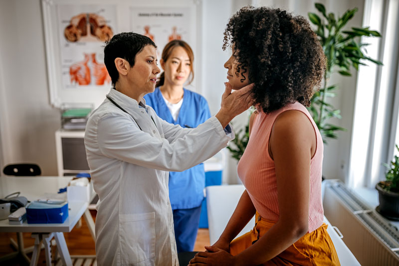A health care provider examines a patient's throat in a medical office, with a nurse observing. The setting is professional and focused, with anatomical charts visible.