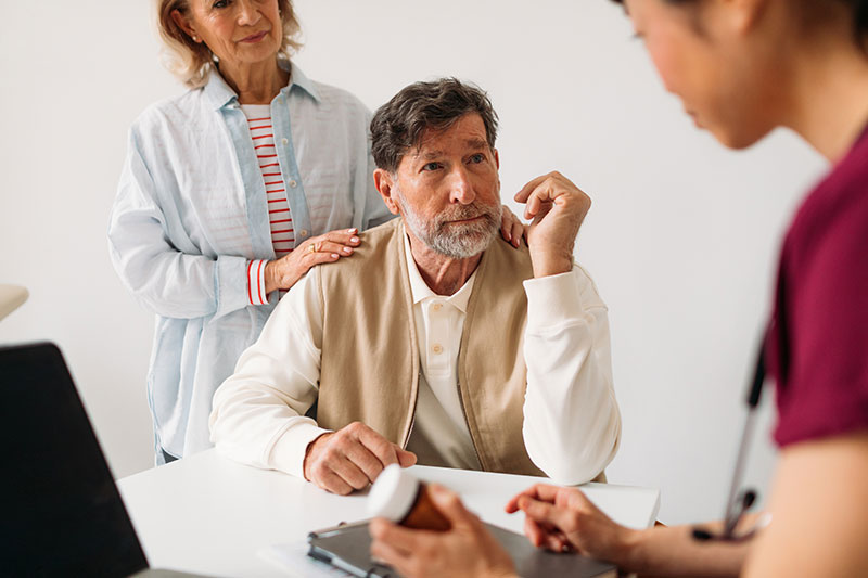 An older man sits at a table, looking thoughtful, with a woman behind him gently resting her hand on his shoulder. A healthcare professional shows him a medication bottle, conveying a caring and supportive atmosphere.