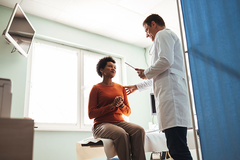 Health care provider in a white coat consults with a patient sitting on an exam table in a bright medical office, conveying a sense of care and attentiveness.