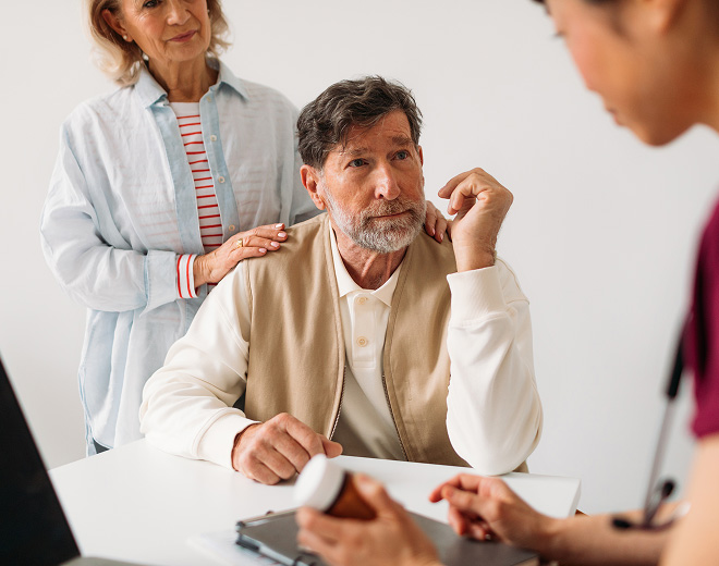 An older man sits at a table, looking thoughtful, with a woman behind him gently resting her hand on his shoulder. A healthcare professional shows him a medication bottle, conveying a caring and supportive atmosphere.