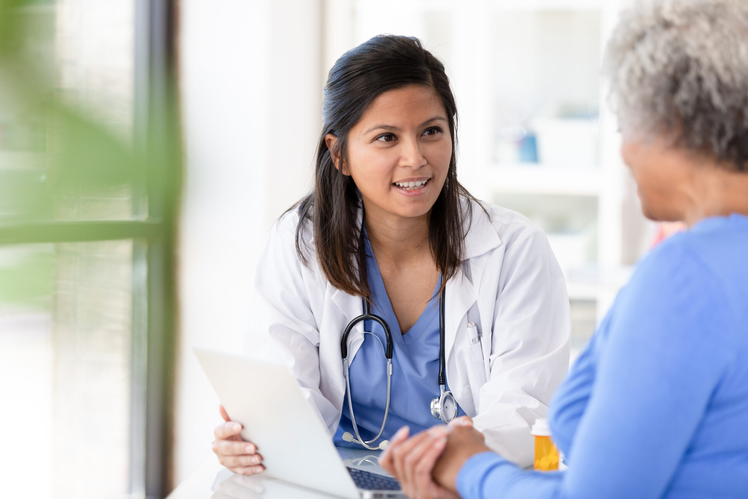 A smiling health care provider in a white coat and stethoscope consults with an older patient, creating a warm, caring atmosphere in a bright medical office.