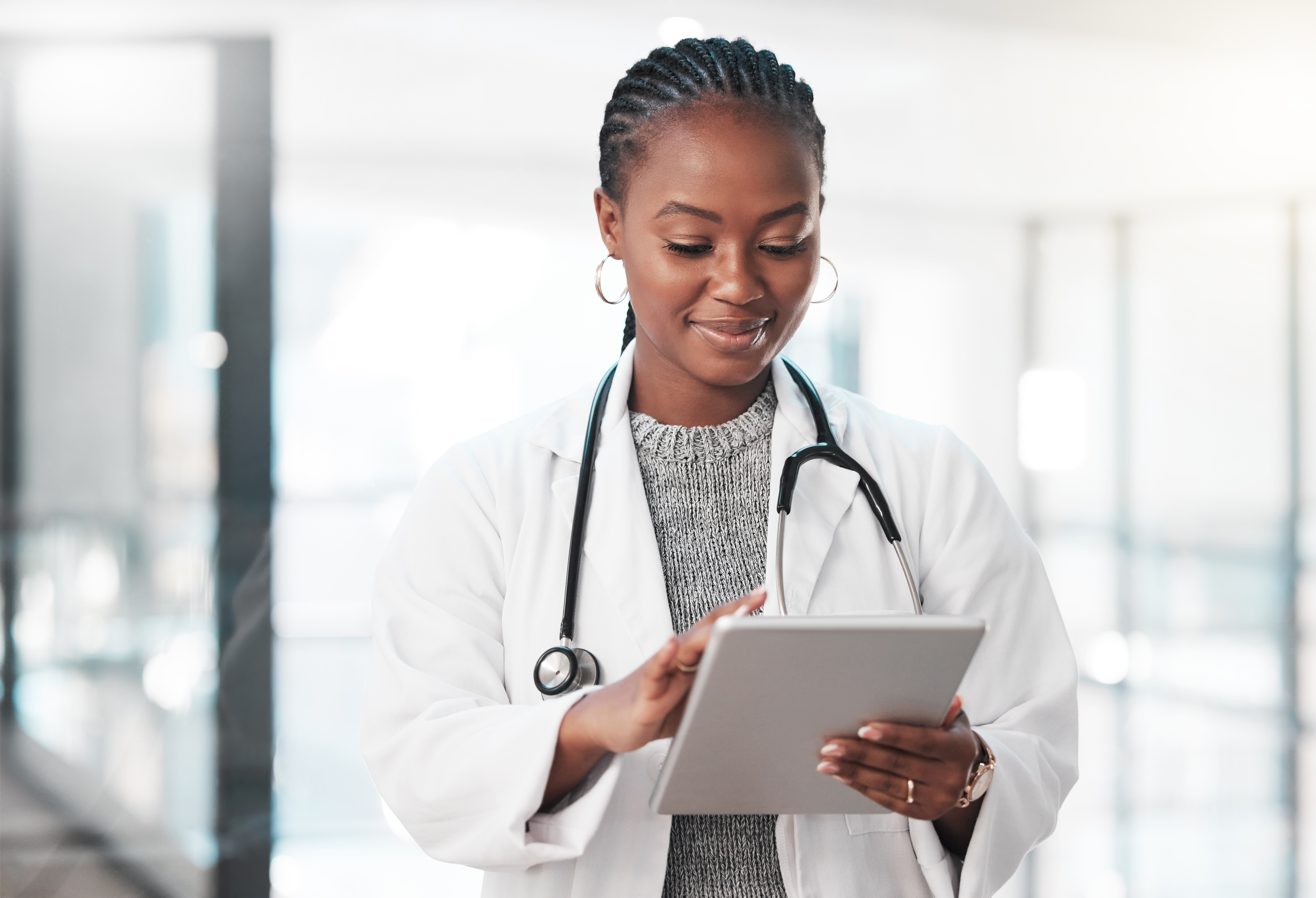 Female health care provider with braided hair and stethoscope uses a tablet in a bright hospital hallway, conveying professionalism and focus.