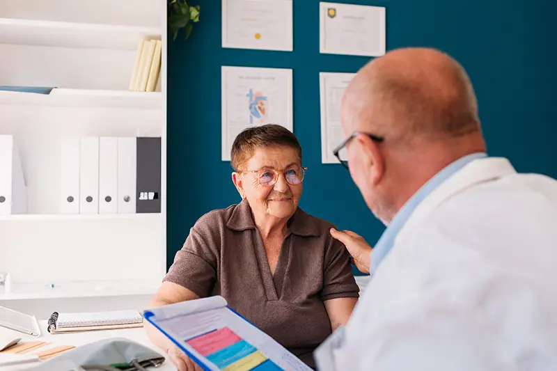 An older woman in a brown shirt sits across from a bald man in a white coat, consulting in a medical office. Both appear engaged and focused.