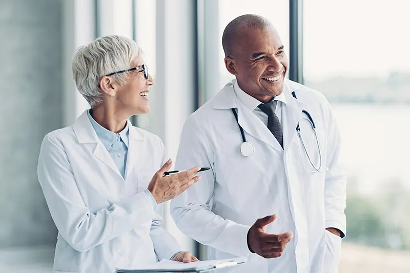 Two health care providers in lab coats share a laugh in a bright hospital hallway. A woman points with a pen, while the man holds his hand up in conversation.