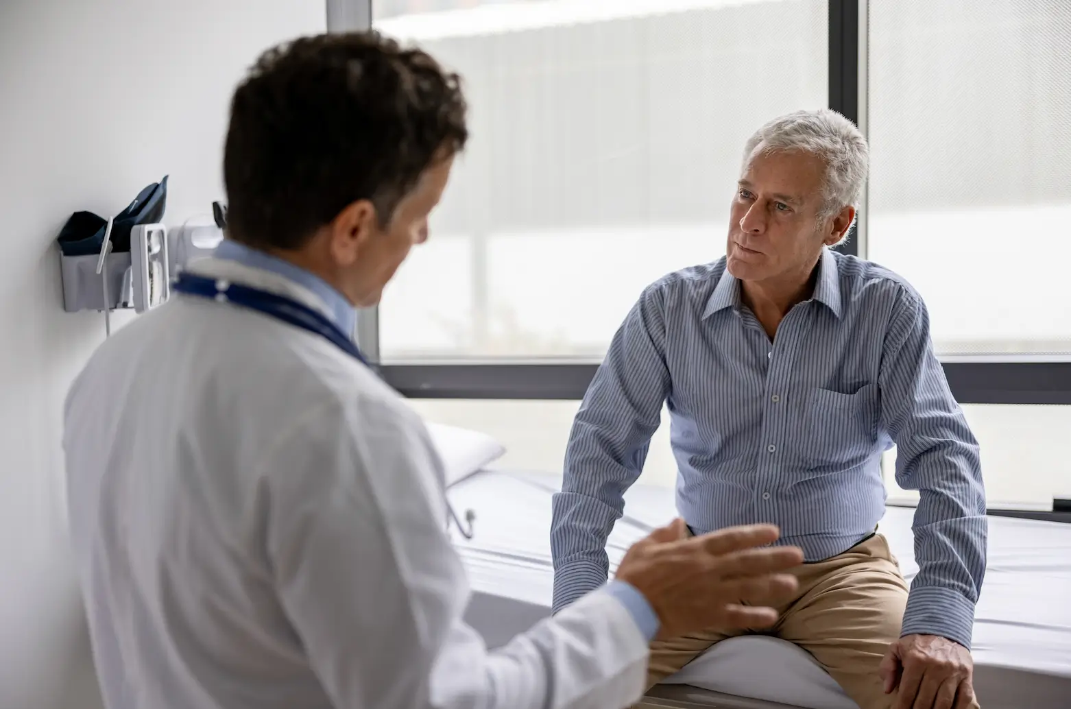 A health care provider in a white coat talks to an older man sitting on an exam table in a medical office. The patient looks attentive and slightly concerned.