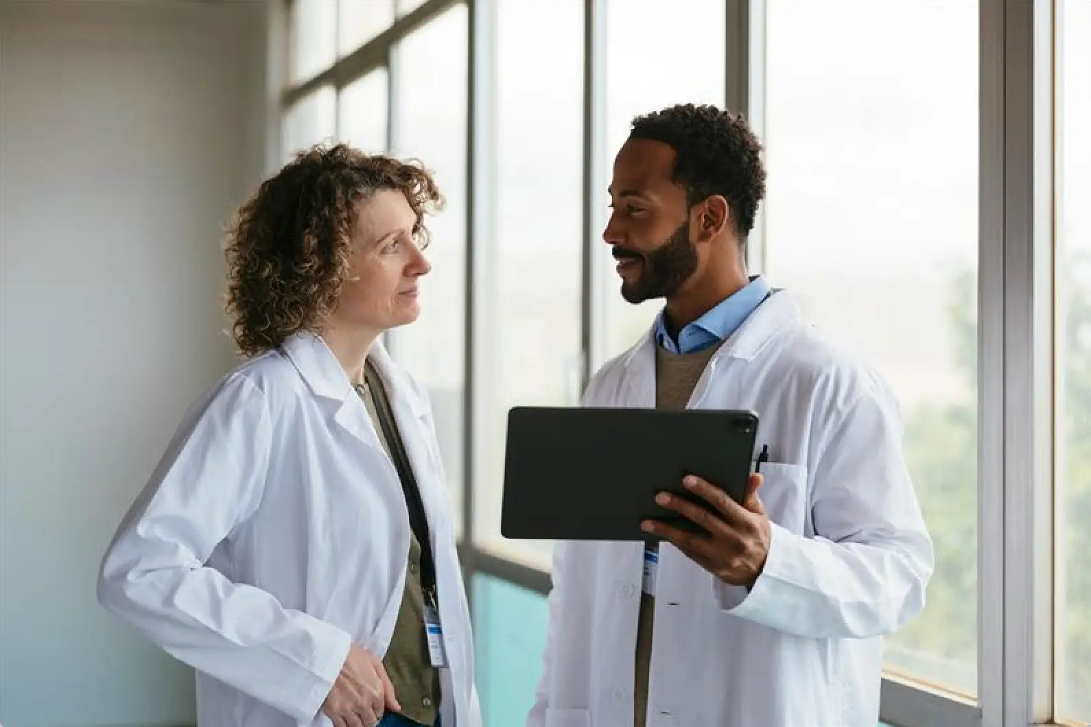 Two health care providers in white coats converse by a sunlit window. One holds a tablet, both smiling, suggesting a positive, collaborative atmosphere.