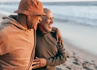 Two friends walking on a beach, smiling and embracing, with the ocean in the background. The scene conveys warmth, companionship, and joy.