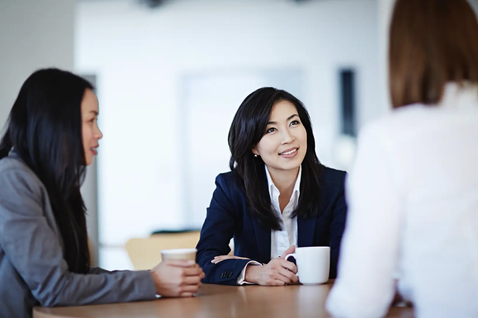 Three people sit at a table during a meeting. A woman in a blue suit smiles, conveying professionalism and engagement, with coffee cups in view.