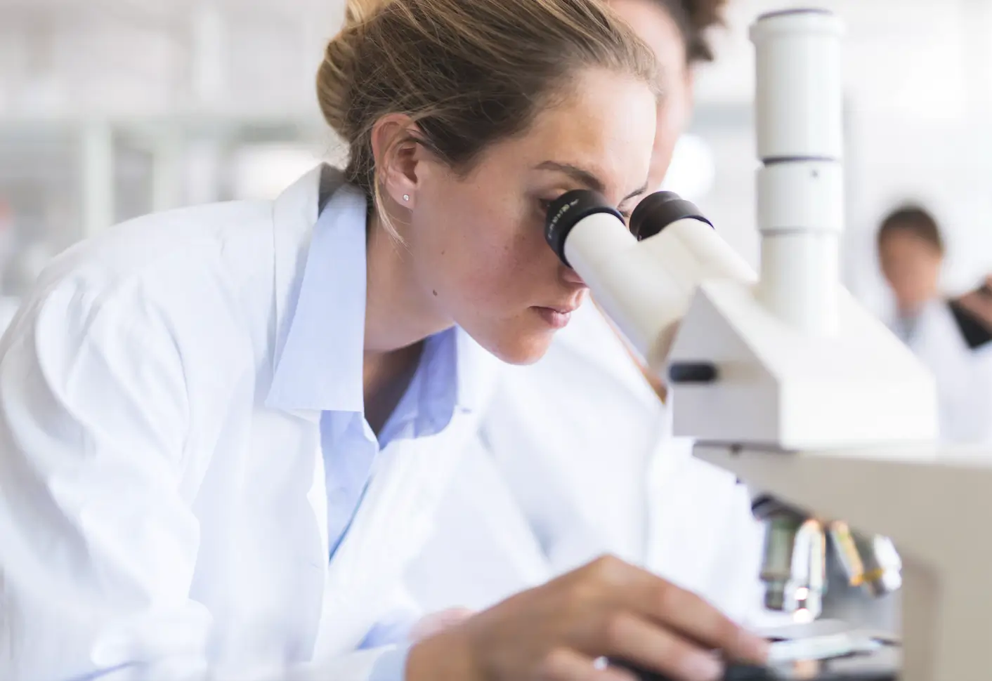 A lab worker in a white lab coat examines a sample through a microscope, focused and determined. The lab setting is bright and clinical.