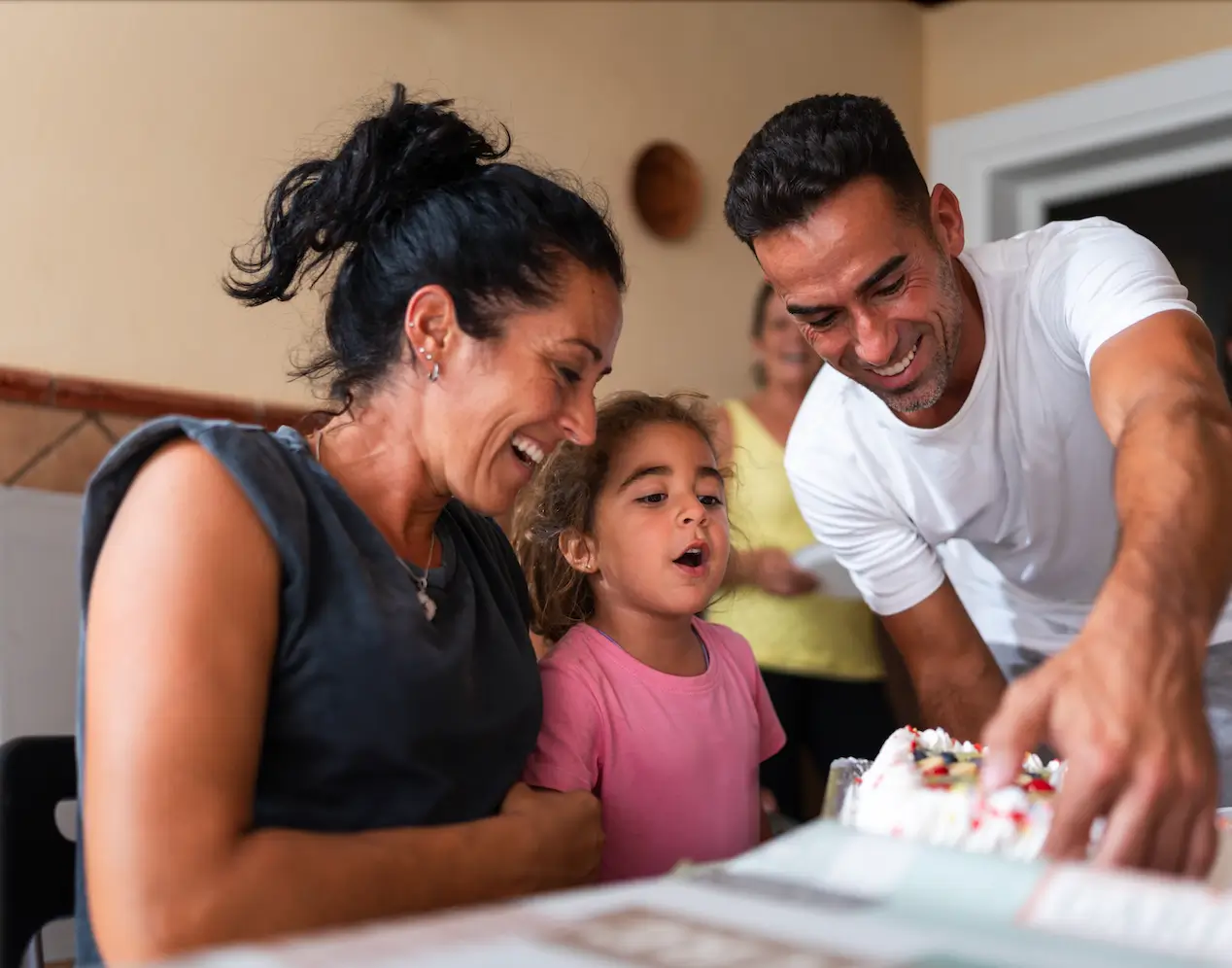 A joyful family moment with a woman and a man smiling as a young girl eagerly blows out candles on a birthday cake, conveying warmth and celebration.