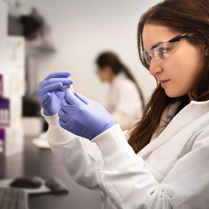 A lab worker in a lab coat and goggles examines a vial closely, wearing purple gloves. Background shows a blurred figure in a lab setting, conveying focus and precision.