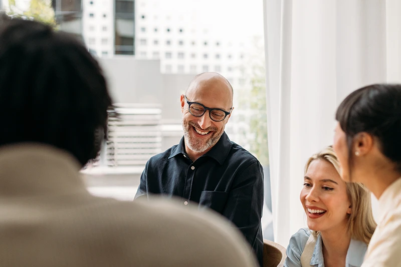 A group of diverse colleagues smiling and chatting in a modern office with large windows. The atmosphere is warm and collaborative.