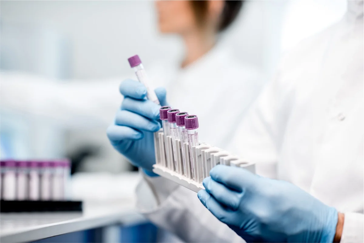 Lab worker in blue gloves carefully handles tray of purple-capped test tubes.