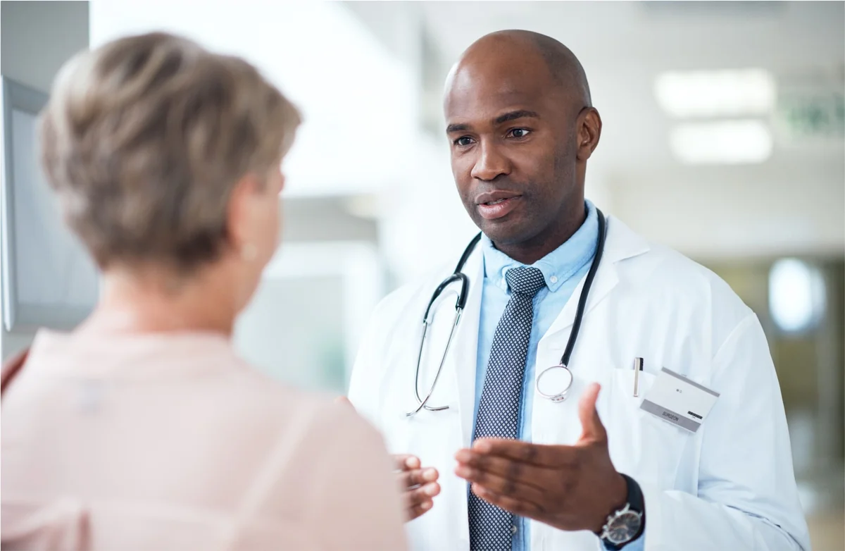 Healthcare provider in a white coat with stethoscope talks to a patient in a bright medical office.