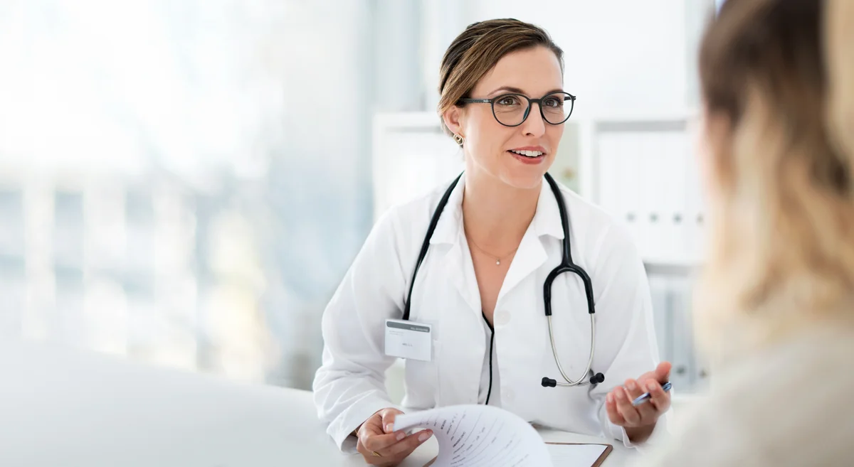 Smiling provider with glasses and stethoscope holds papers and talks to patient in bright office.