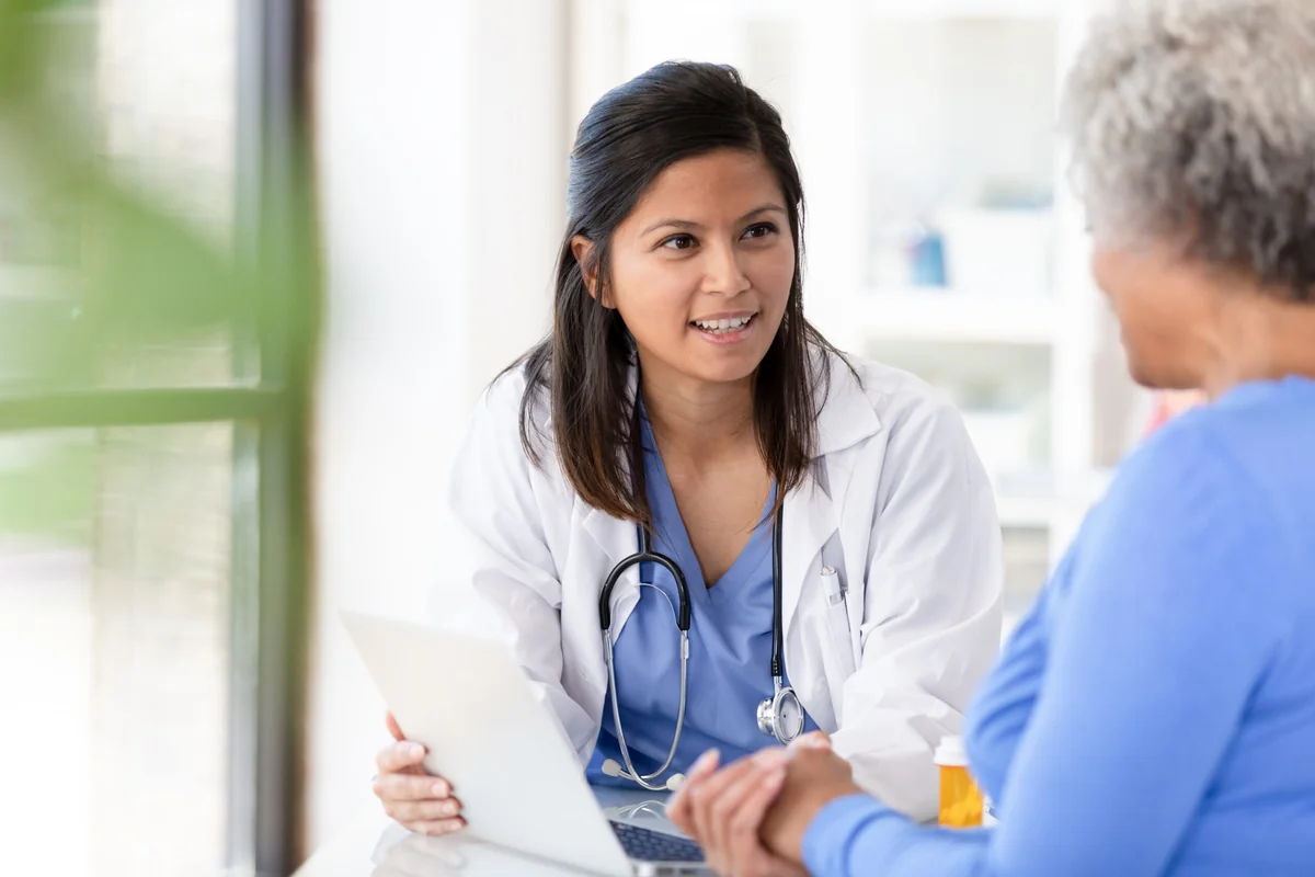 Smiling provider in white coat and stethoscope consults older patient in a warm, bright office.